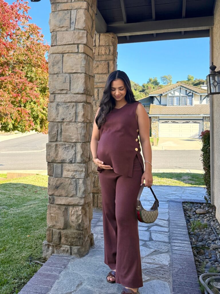 Pregnant woman in a brown sleeveless knit tank and matching wide-leg knit pants with a small logo bag in front of a house with autumn foliage