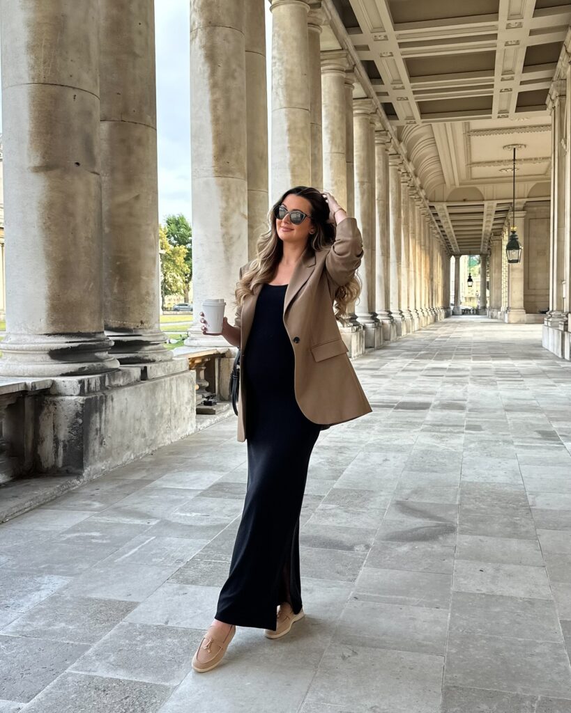 Pregnant woman in a camel blazer over a black bodycon maxi dress with tan suede loafers holding a coffee in a stone colonnade