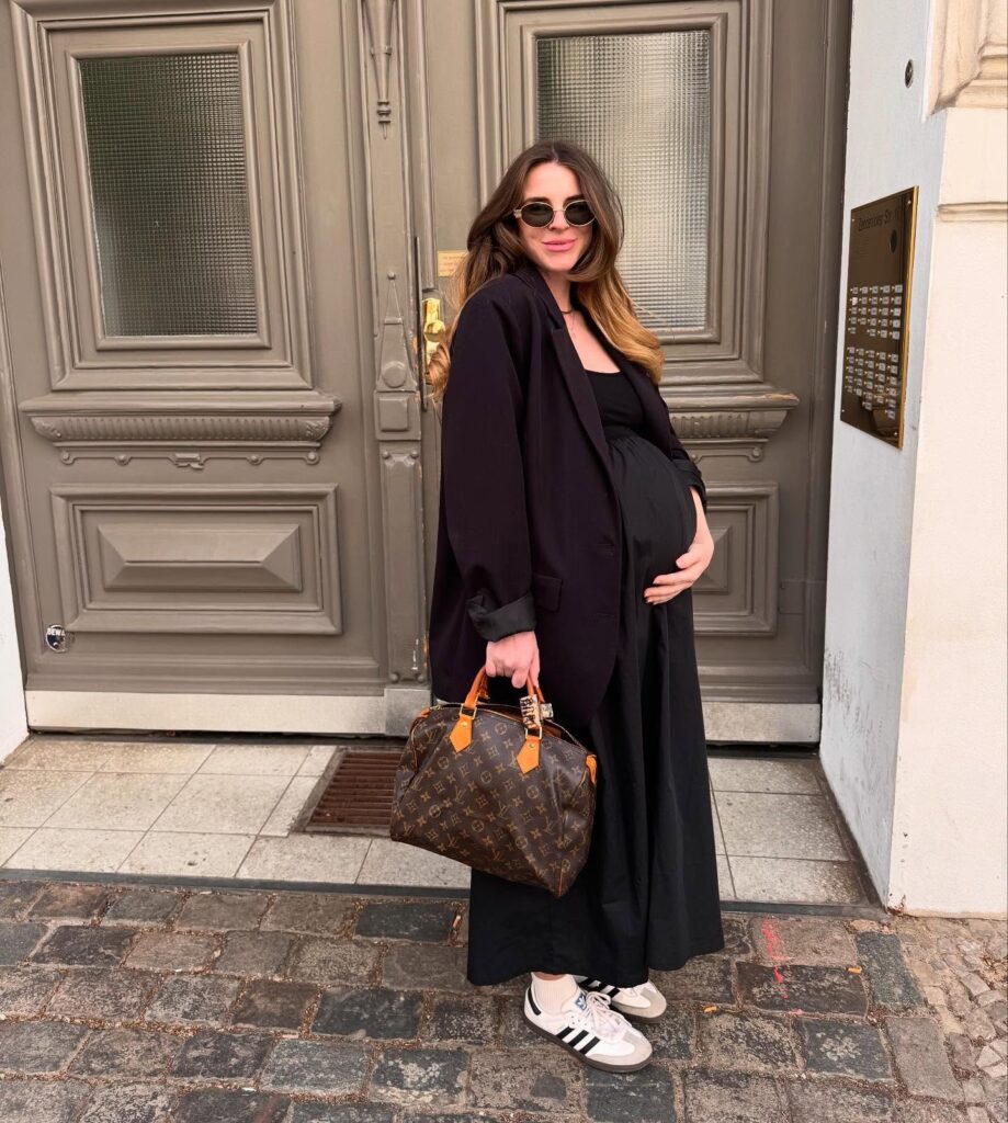 Pregnant woman in an oversized black blazer over a black knit maxi dress with white Adidas Samba sneakers and a monogram bag