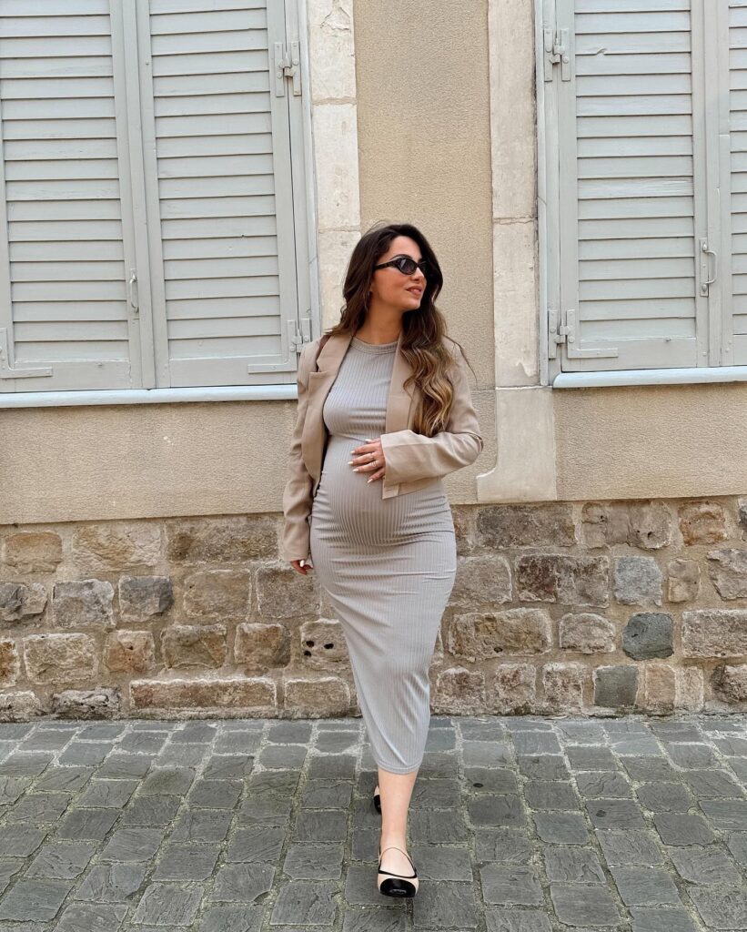 Pregnant woman in a beige ribbed bodycon midi dress with a tan blazer over her shoulders and cap-toe flats on a cobblestone street