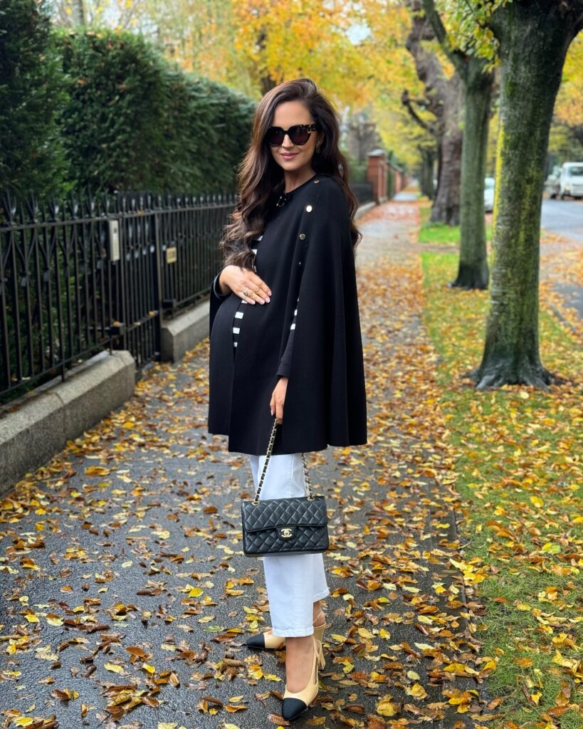 Pregnant woman in a black wool cape over white wide-leg jeans with cap-toe slingbacks and a black quilted chain bag on a leaf-covered street