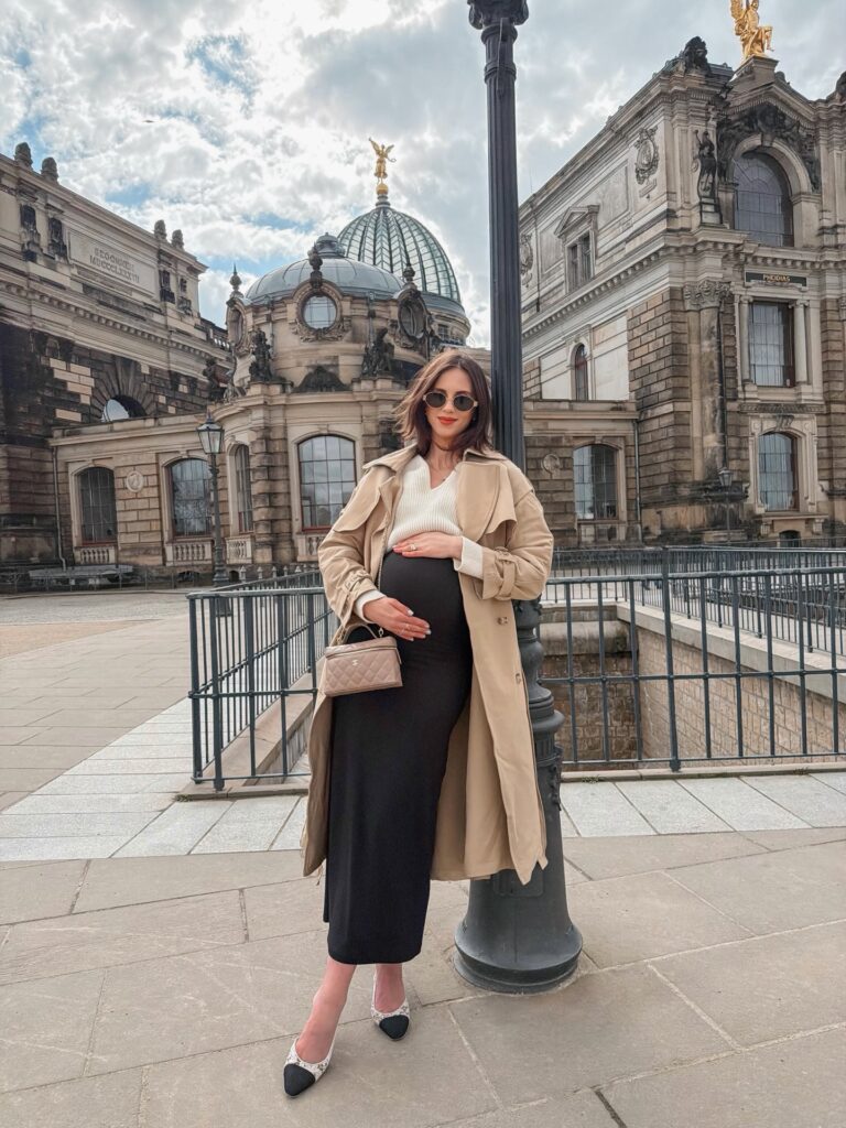 Pregnant woman in a beige classic trench coat, cream V-neck sweater and black knit maxi skirt with cap-toe slingback flats