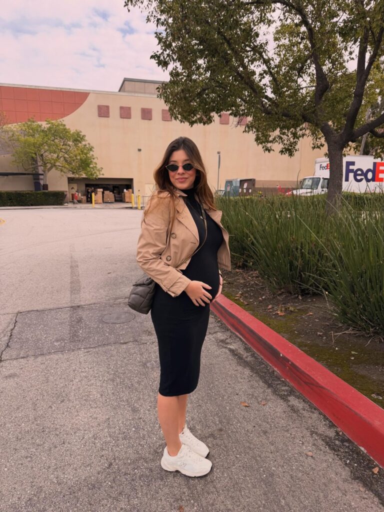 Pregnant woman in a cropped tan trench jacket over a black ribbed bodycon midi dress with white chunky sneakers