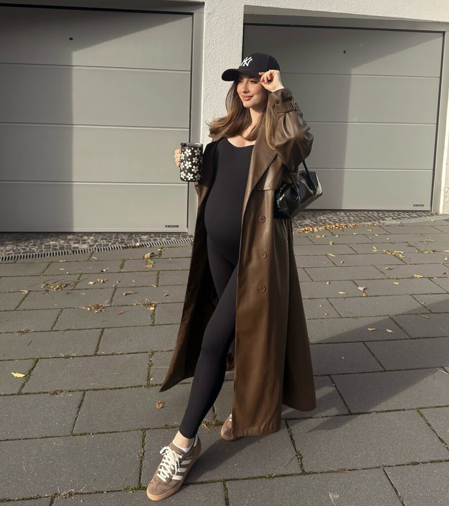 Pregnant woman in a long brown faux-leather trench coat over a black unitard with a navy baseball cap and brown suede Adidas Samba sneakers