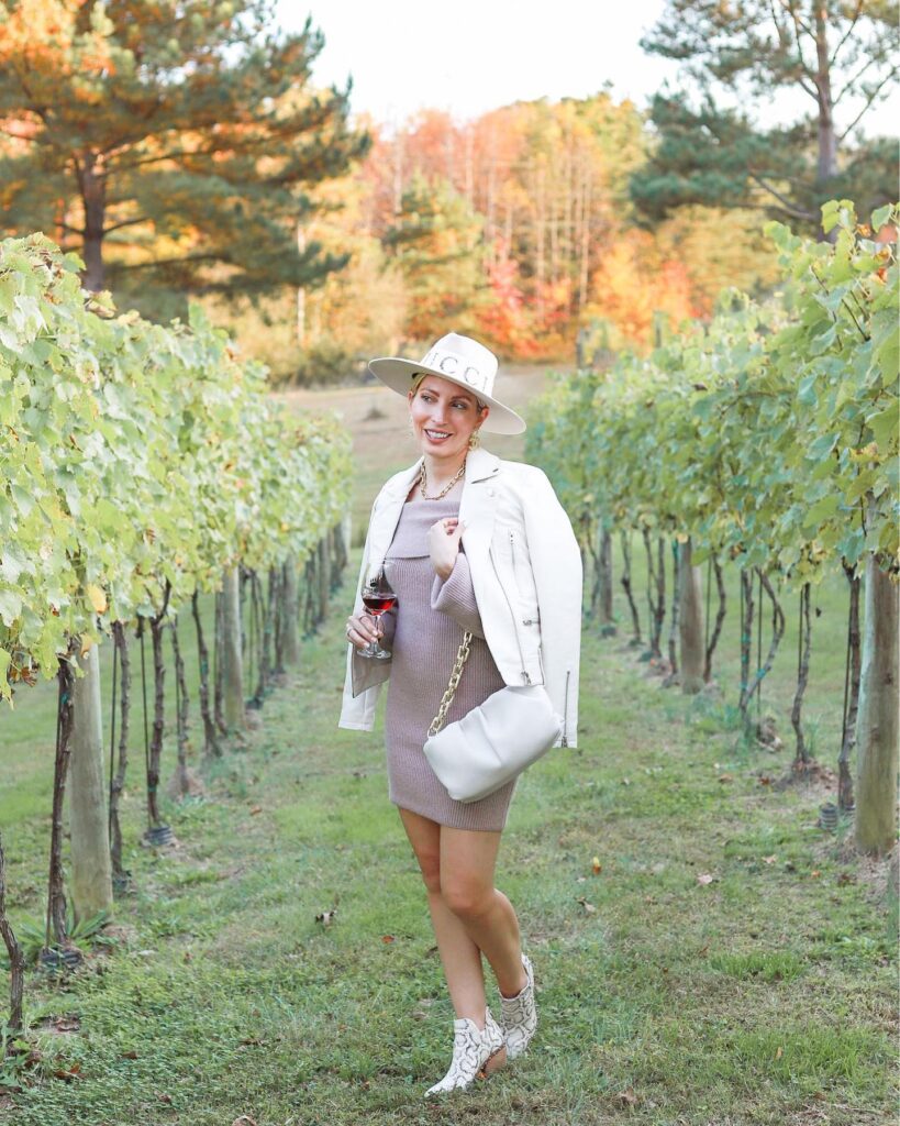 Woman in a taupe sweater dress, white moto jacket and wide-brim hat with snakeskin boots at a vineyard