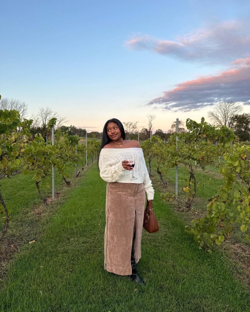 Woman in a cream off-shoulder cable knit sweater and tan slip skirt at sunset in a vineyard