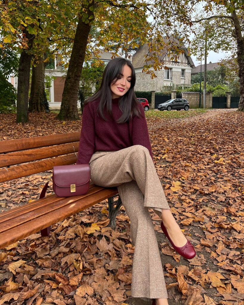 Woman in a burgundy sweater and camel tweed flare trousers sitting on a bench among autumn leaves