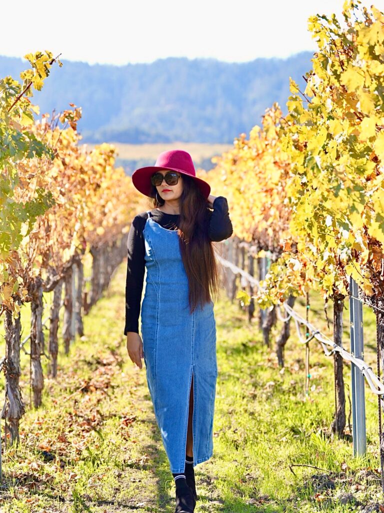 Woman in a denim slip midi dress over a black long-sleeve tee with a berry felt hat in a golden vineyard