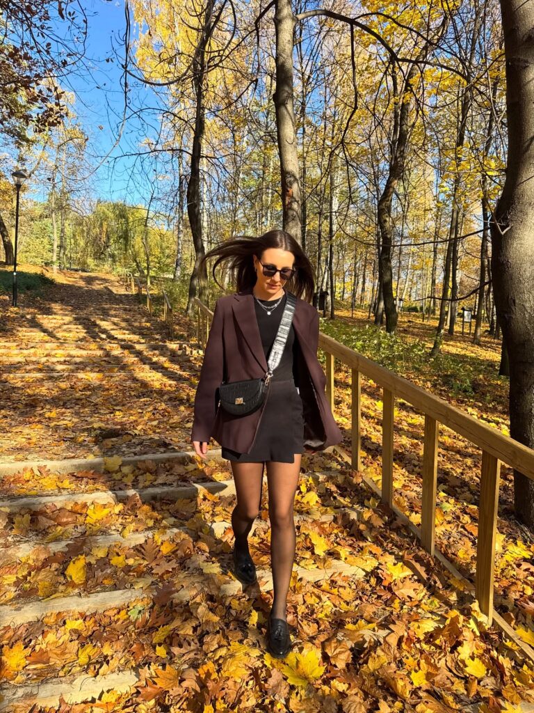 Woman in a brown blazer, black tee, black skort, sheer tights and loafers walking down leaf-covered stairs