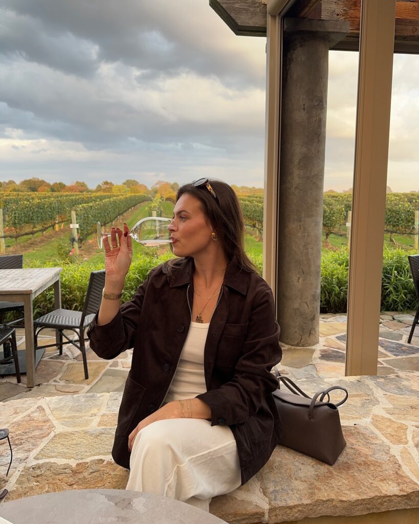 Woman in a brown suede shirt jacket over a cream tank and cream wide pants on a vineyard patio at sunset