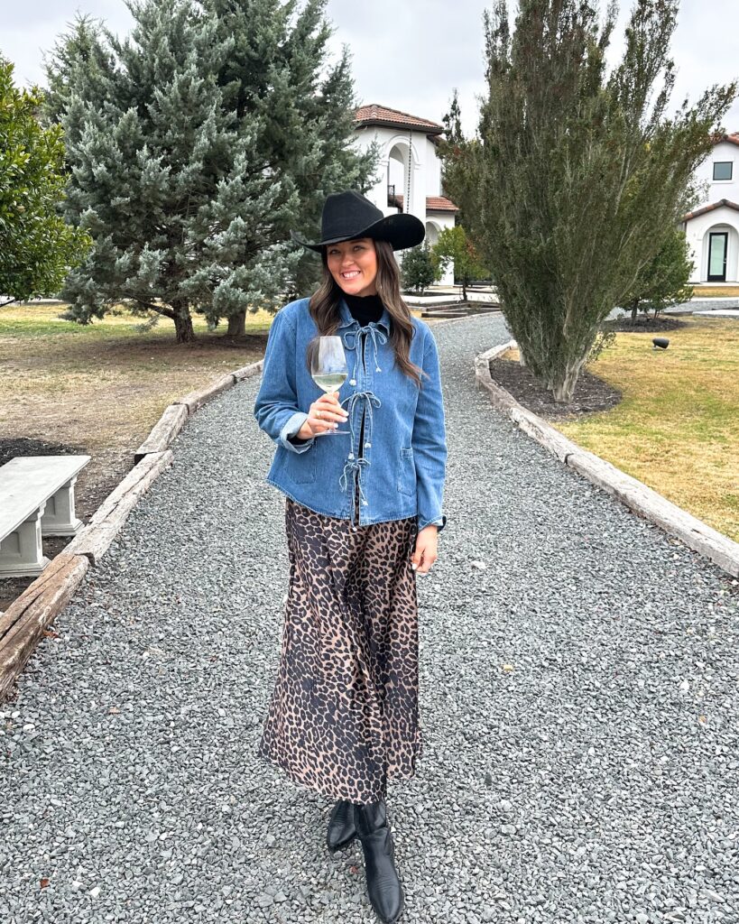 Woman in a denim jacket, black turtleneck, leopard satin maxi skirt, black cowboy hat and boots
