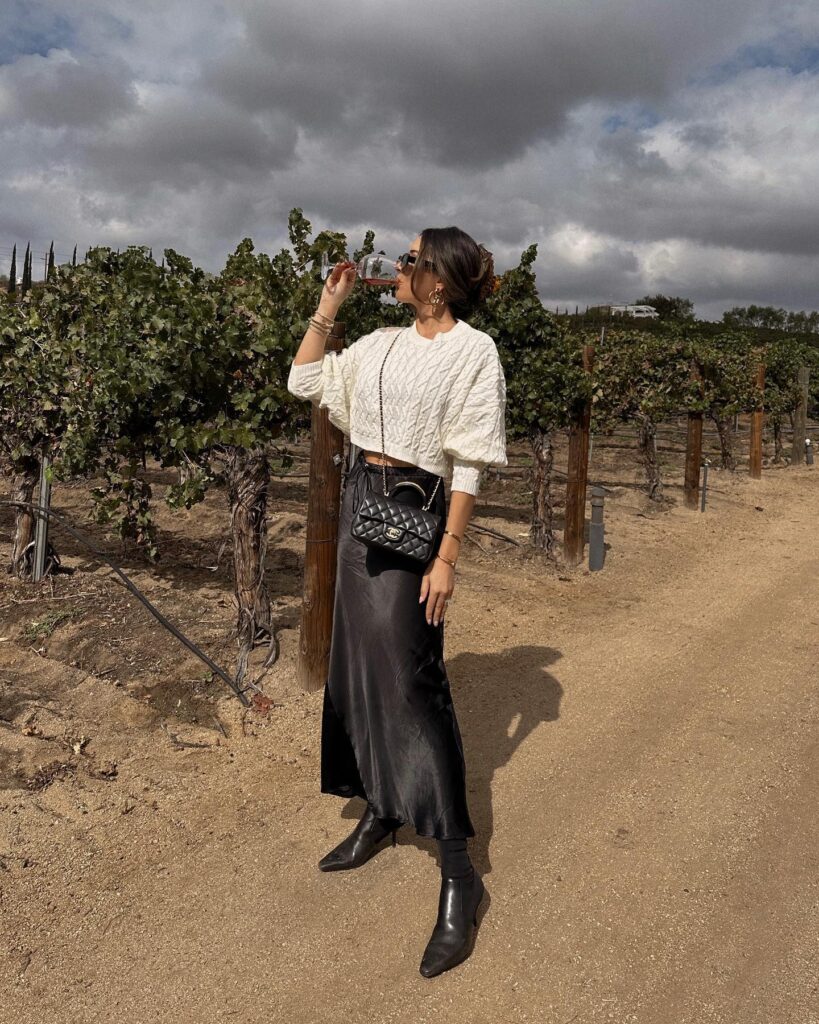 Woman in a cream cropped cable sweater and black satin slip skirt with black booties in a vineyard