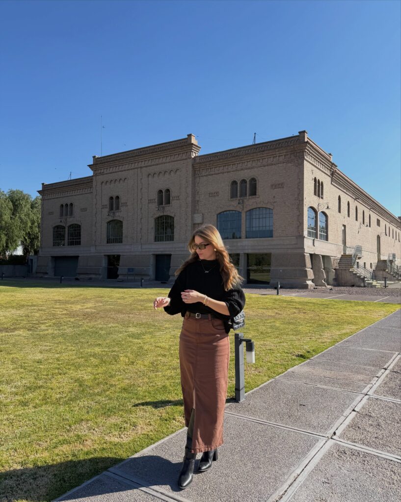 Woman in a black sweater, brown denim maxi skirt and black boots in front of a brick building