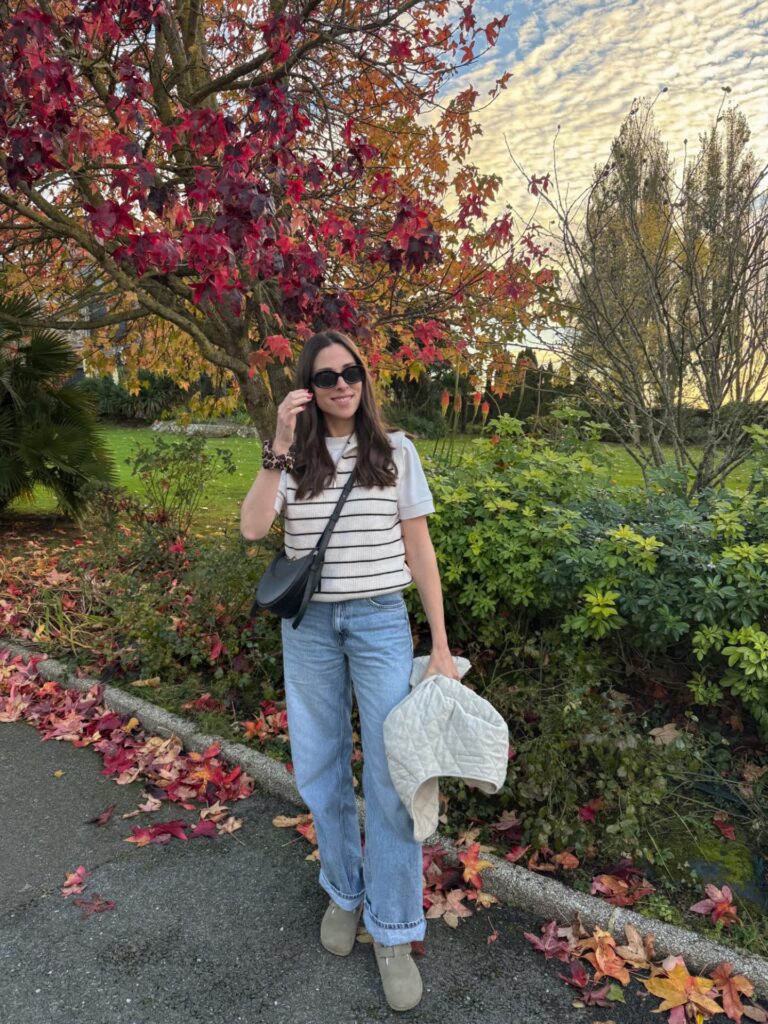 Woman in a striped sweater vest over a tee, light wide-leg jeans and suede clogs with a quilted jacket in hand