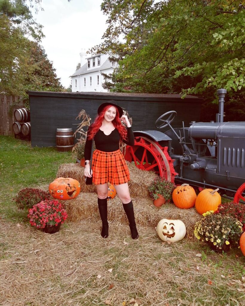 Woman in a black top, orange plaid mini skirt, black hat and over-the-knee boots near pumpkins and a tractor