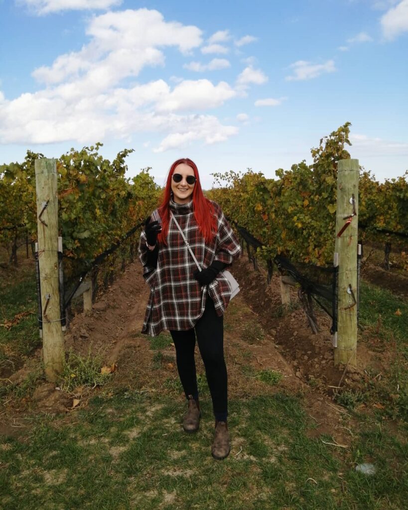 Woman in a red and black plaid poncho, black leggings and Chelsea boots between rows of vines
