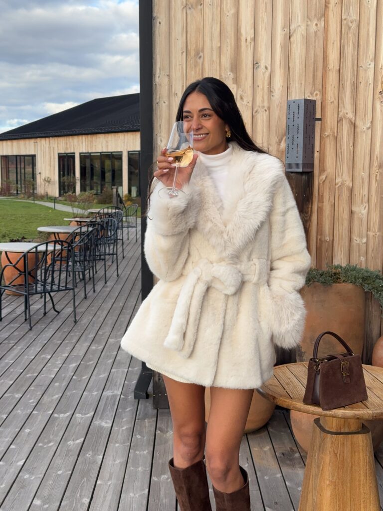 Woman in a cream faux-fur coat over a white turtleneck with brown tall boots on a winery deck