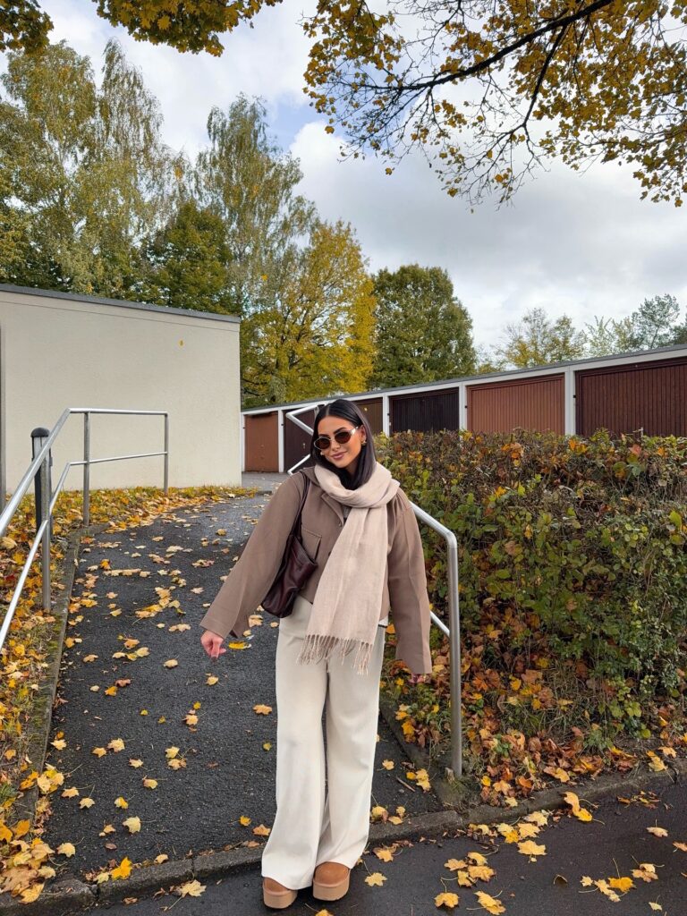 Woman in a taupe cropped jacket, oversized beige scarf, cream wide pants and platform boots among fall leaves