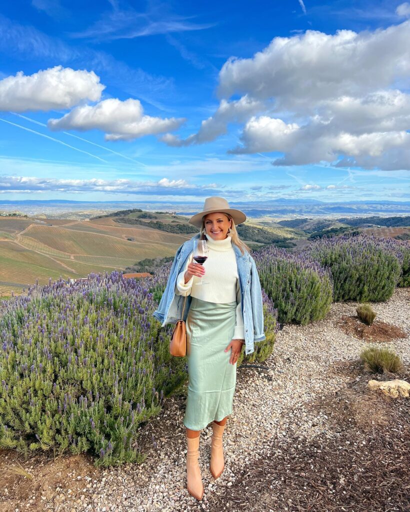Woman in a cream turtleneck, sage slip skirt, denim jacket and felt hat holding red wine