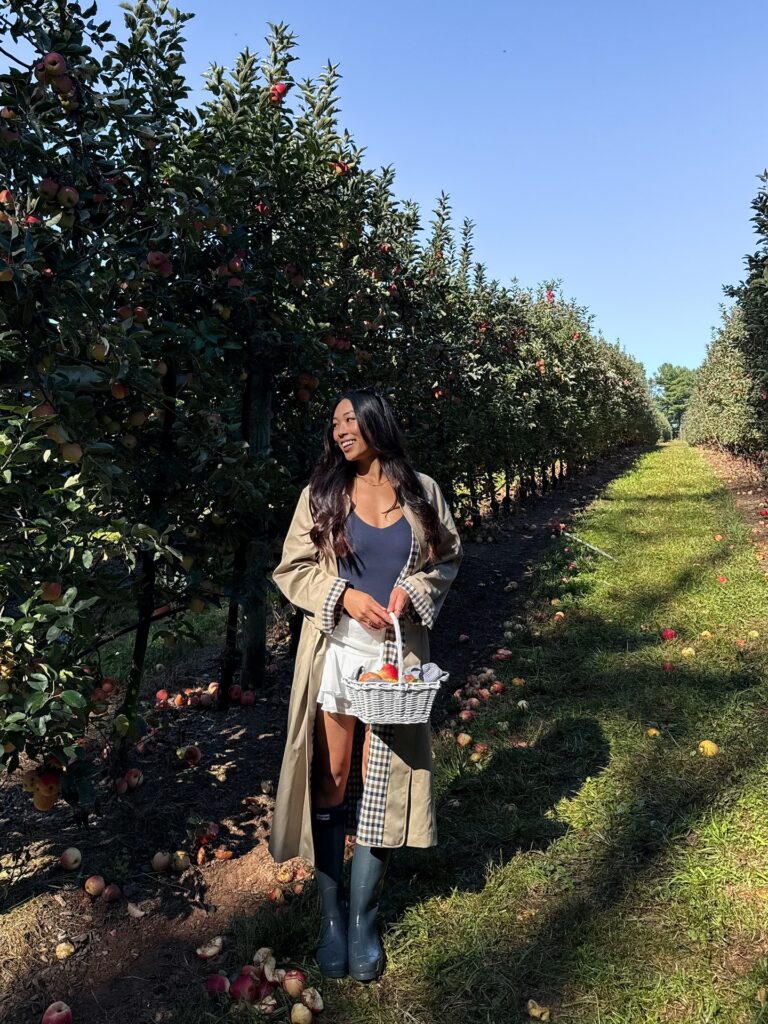 Woman in a navy bodysuit, white skort, long trench coat and Hunter rain boots in an apple orchard