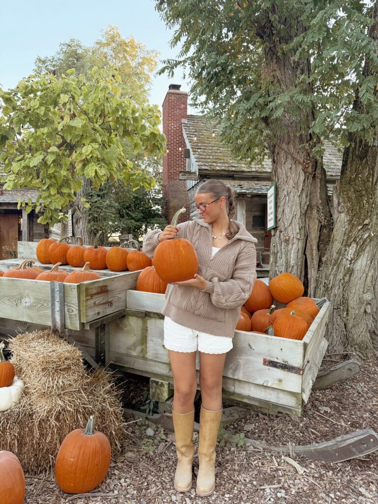 Woman in a taupe cable quarter-zip sweater, white eyelet shorts and tan tall boots at a pumpkin patch