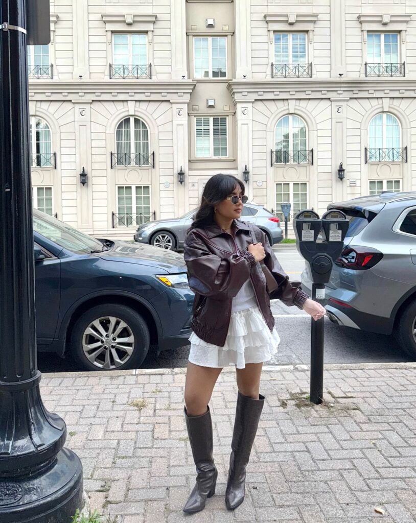 Brown leather bomber jacket over a white tiered ruffle mini skirt with black knee-high boots