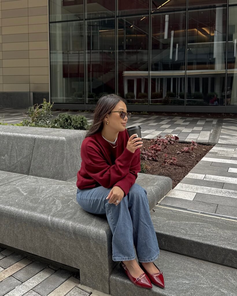 Burgundy crewneck sweatshirt with straight-leg jeans and red patent pointed-toe flats, holding a coffee