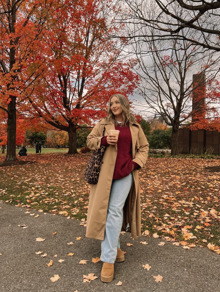 Camel trench coat over a burgundy sweater with light-wash jeans, UGG mini boots and a leopard-print tote among autumn leaves