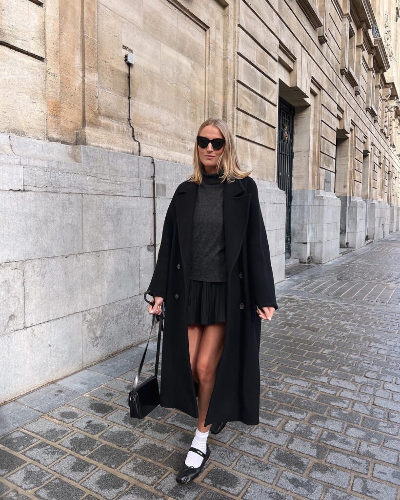 Long black wool coat over a grey sweater and dark pleated mini skirt with black ballet flats and white socks