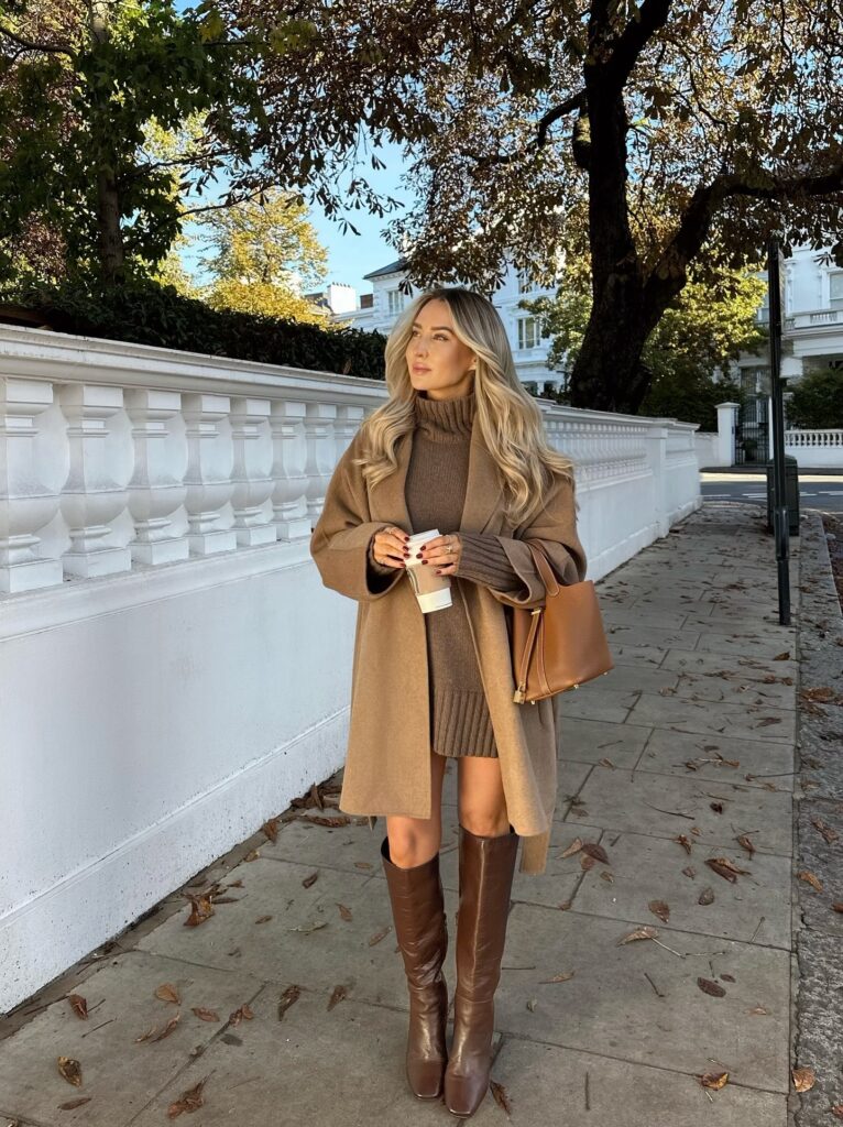 Woman in a camel wool coat over a brown roll-neck knit dress with brown leather knee boots and a tan bag among autumn leaves