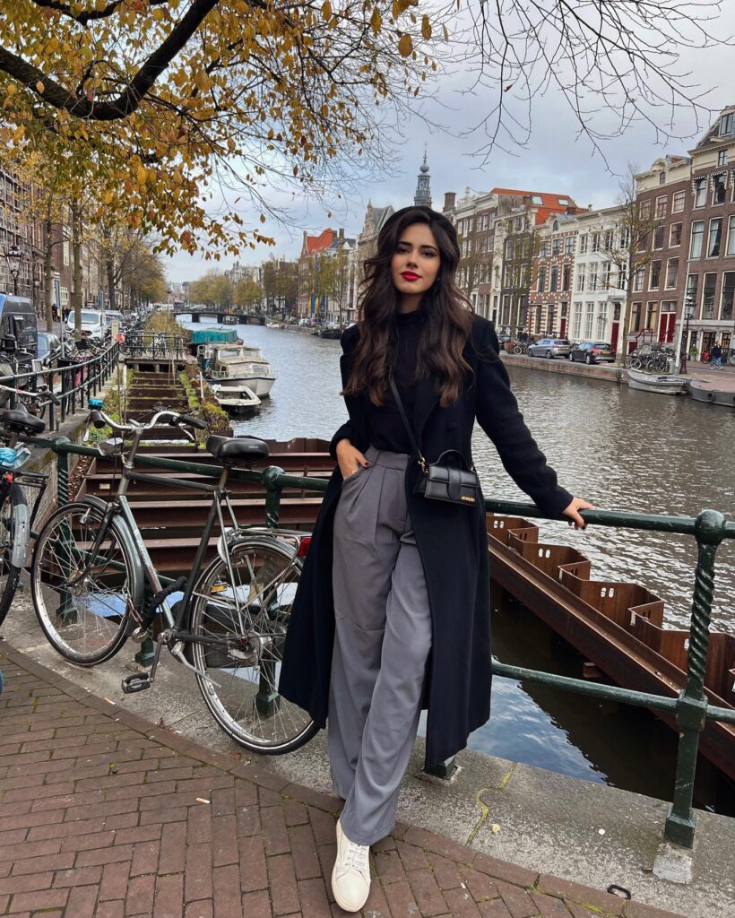 Woman in a long black tailored coat over a black top with grey pleated wide-leg trousers, a small bag and white sneakers by a canal