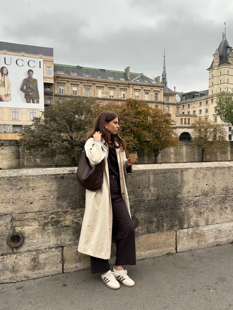 Woman in a cream trench coat over a brown top and brown trousers with a suede bag and Adidas Samba sneakers