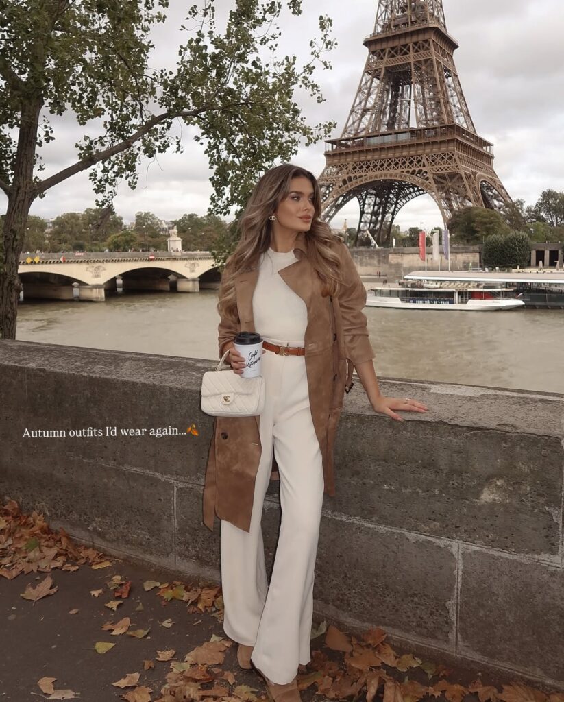 Woman in a tan suede trench coat over a cream knit and matching cream wide-leg trousers with a quilted flap bag by the Seine