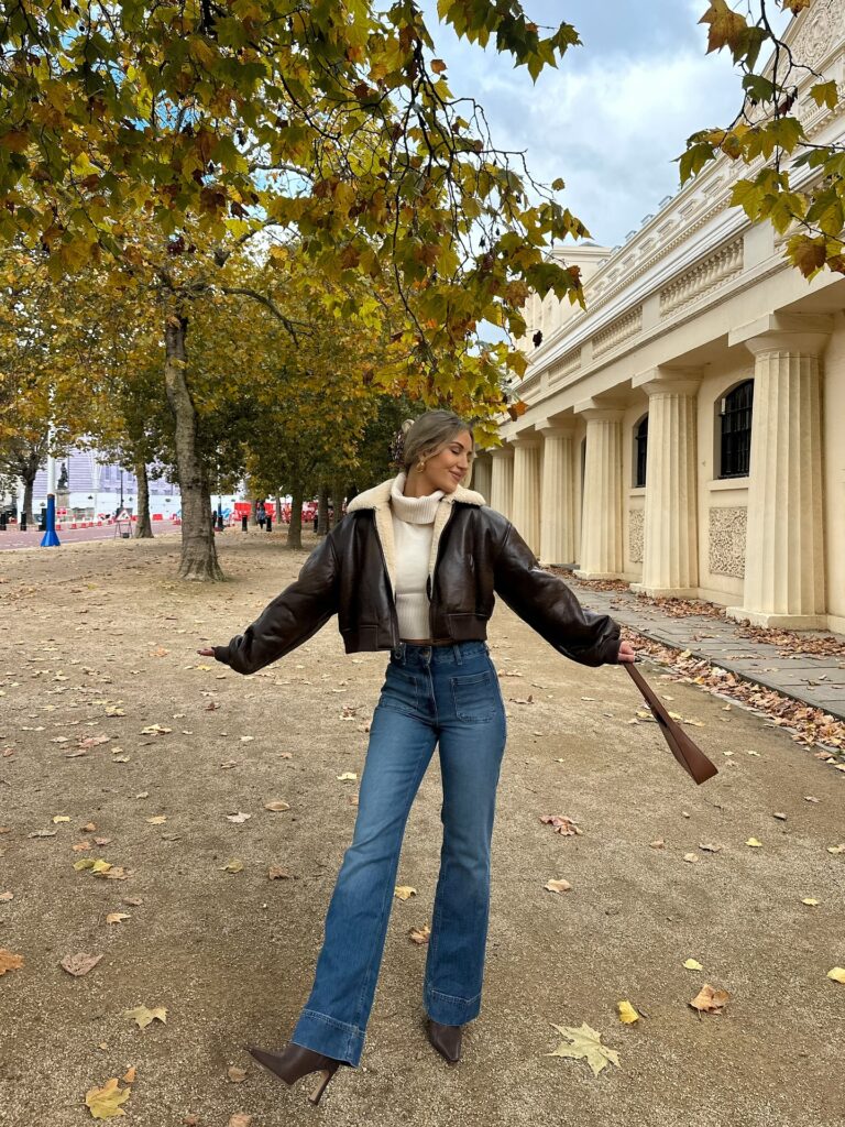 Woman in a brown leather jacket with a shearling collar over a cream roll-neck with blue flared jeans and brown boots among autumn leaves
