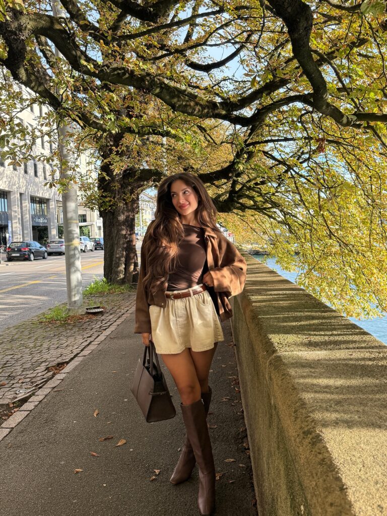 Woman in a brown suede shirt-jacket over a fitted top with a cream skirt and tall brown knee boots by a lake