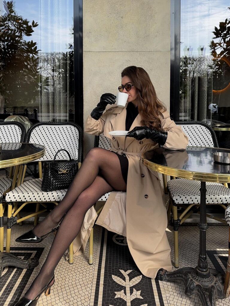 Woman seated at a Parisian café in a beige trench coat with black leather gloves, sheer tights and black patent slingbacks