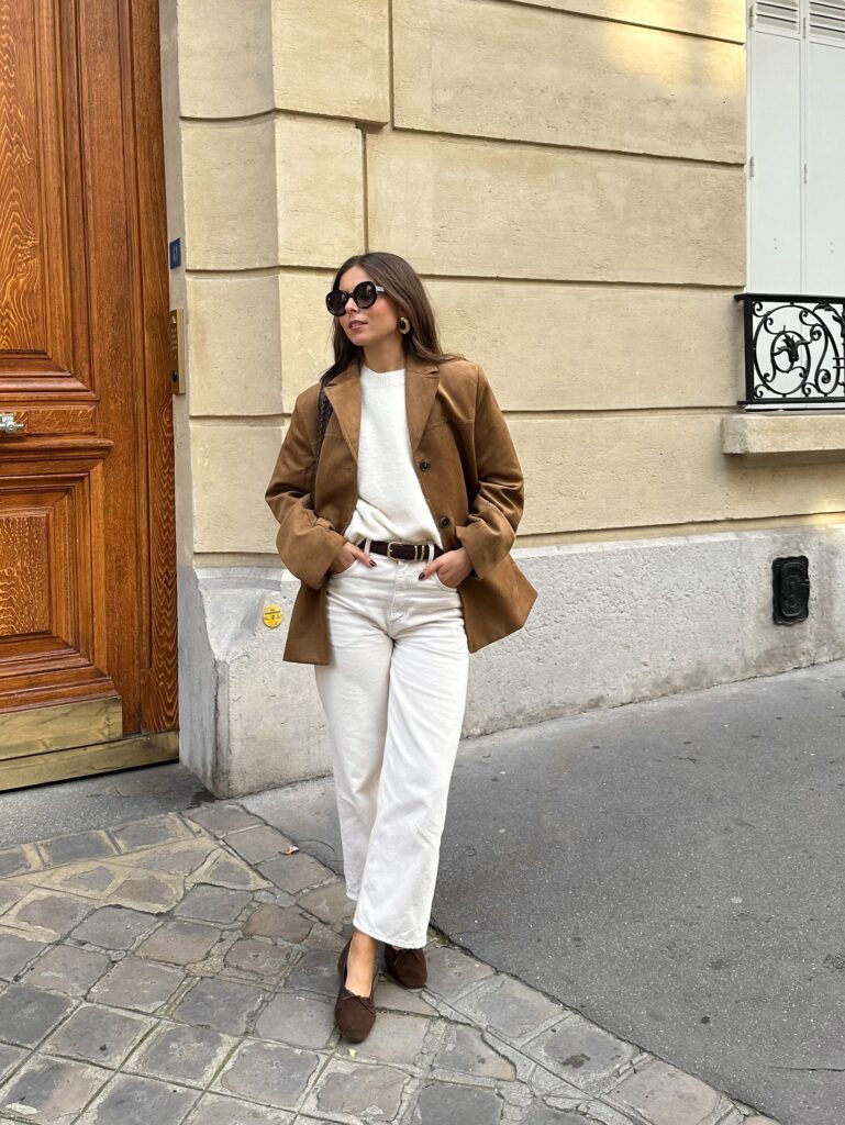 Woman in a tan suede blazer over a cream knit with ecru straight-leg jeans and brown flats on a Paris street