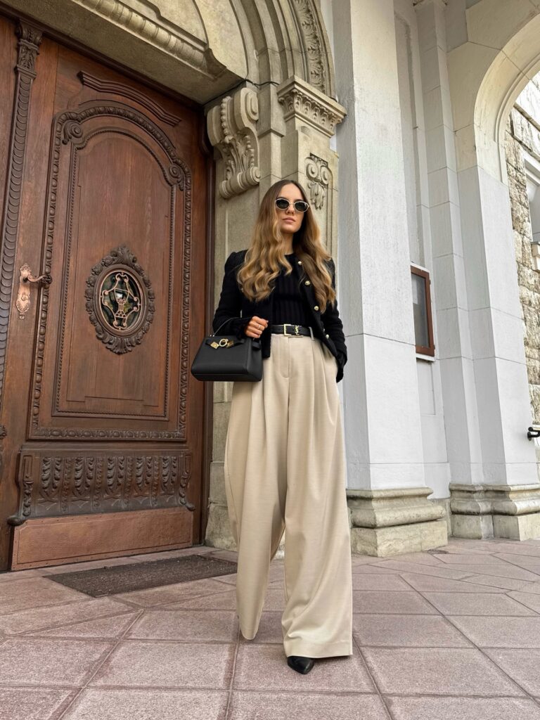 Woman in a black tweed jacket with gold buttons over a black top with beige wide-leg tailored trousers and a black top-handle bag