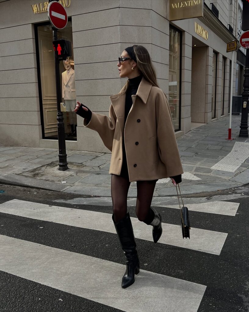 Woman in a camel wool coat over a brown roll-neck knit dress with brown leather knee boots and a tan bag among autumn leaves