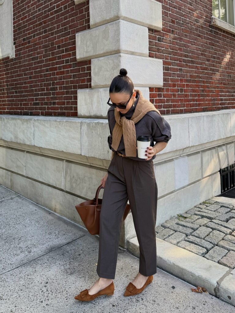 Woman in a charcoal puff-sleeve jumper with a camel cable-knit scarf, brown tailored trousers, suede buckle loafers and a brown tote