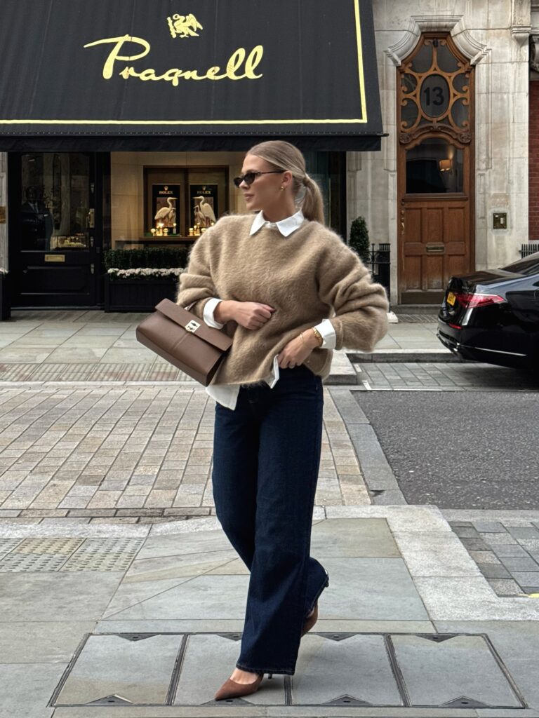 Woman in a camel mohair jumper layered over a white collared shirt with dark indigo wide-leg jeans, brown heels and a brown top-handle bag