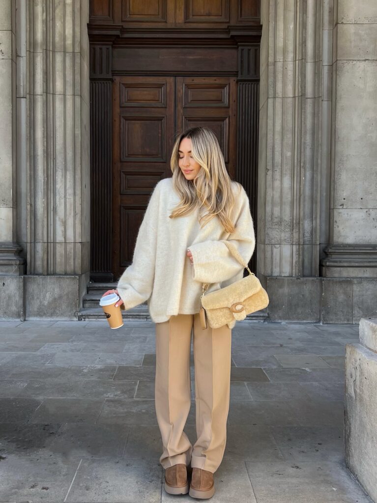 Woman in a cream oversized fuzzy jumper with camel trousers, a cream shearling bag and tan mini boots by stone columns