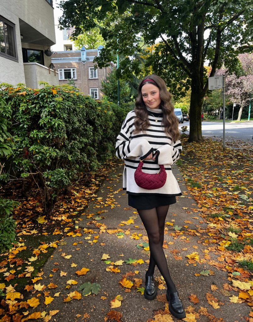 Woman in a black-and-white striped roll-neck jumper over a black mini with sheer tights, chunky black loafers and a burgundy bag among autumn leaves