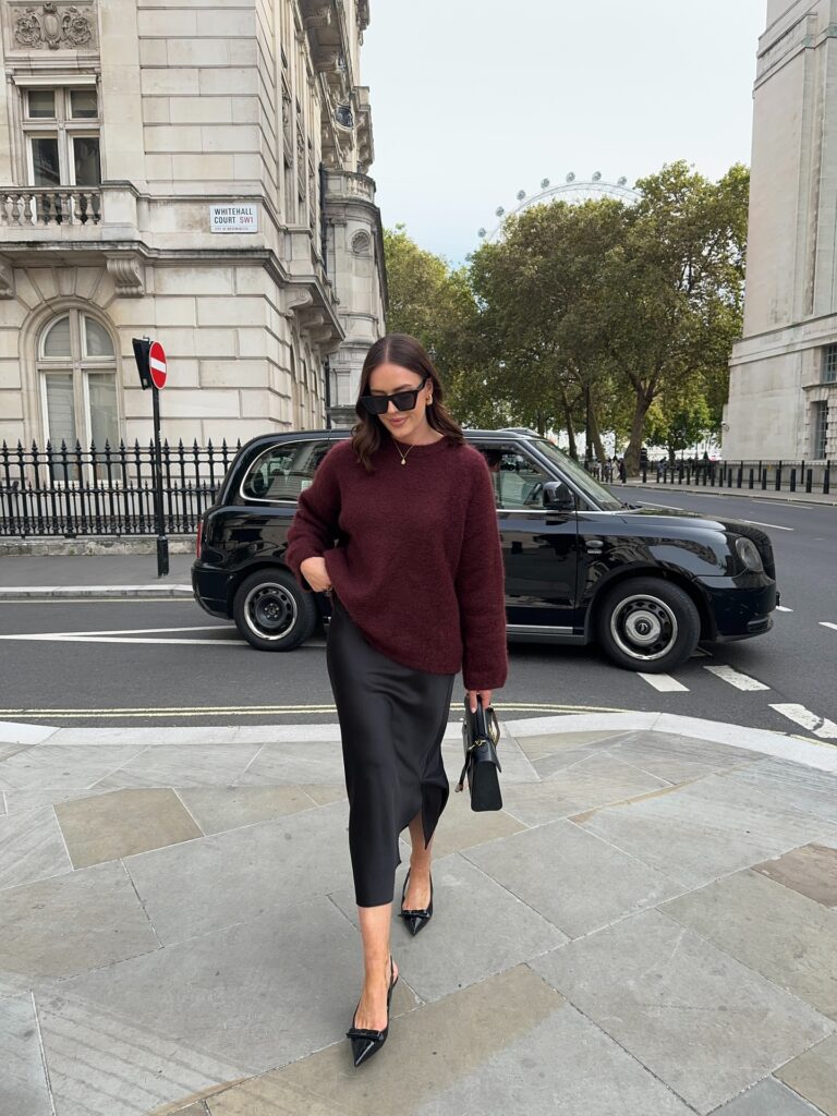 Woman in a burgundy mohair jumper with a black satin midi skirt and black pointed slingback heels on a London street