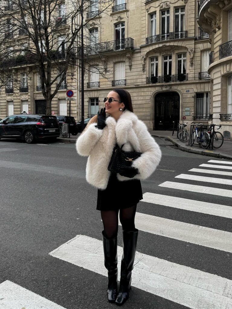 Woman crossing a Paris street in a cream fur jacket over a black mini skirt with black tights and tall black knee boots
