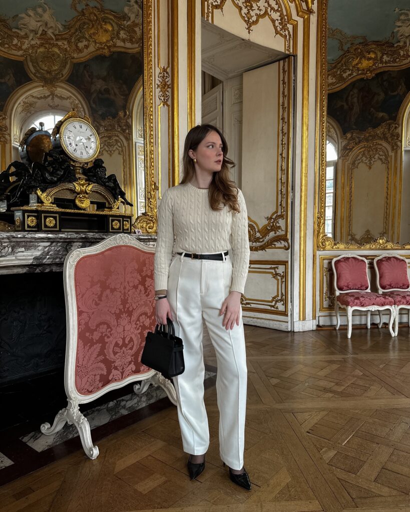 Woman in a cream cable-knit jumper tucked into ivory tailored trousers with a black belt and pointed heels in an ornate gilded room