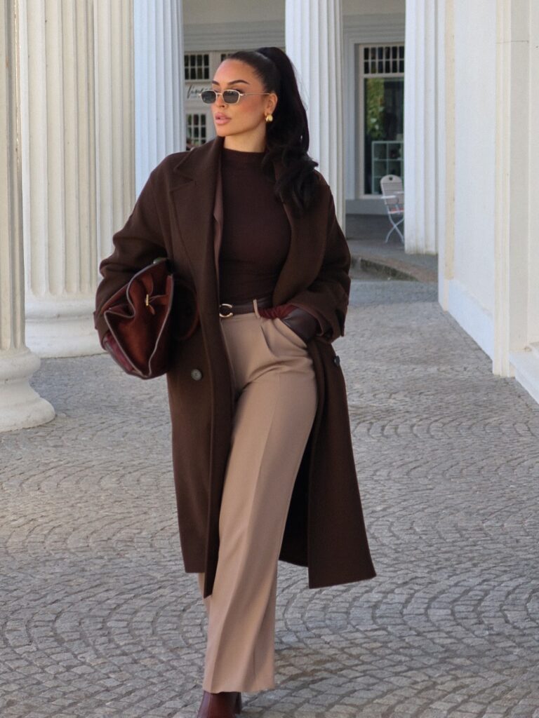 Woman in a long brown wool coat over a brown roll-neck with taupe tailored trousers and an oxblood bag by white columns