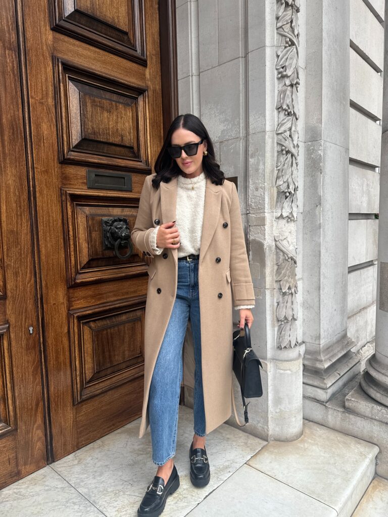 Woman in a tailored camel double-breasted coat over a cream knit with blue jeans and black loafers