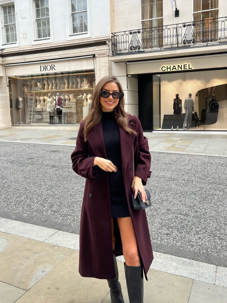 Woman in a long burgundy wool coat over a black knit dress with black knee boots in front of designer storefronts