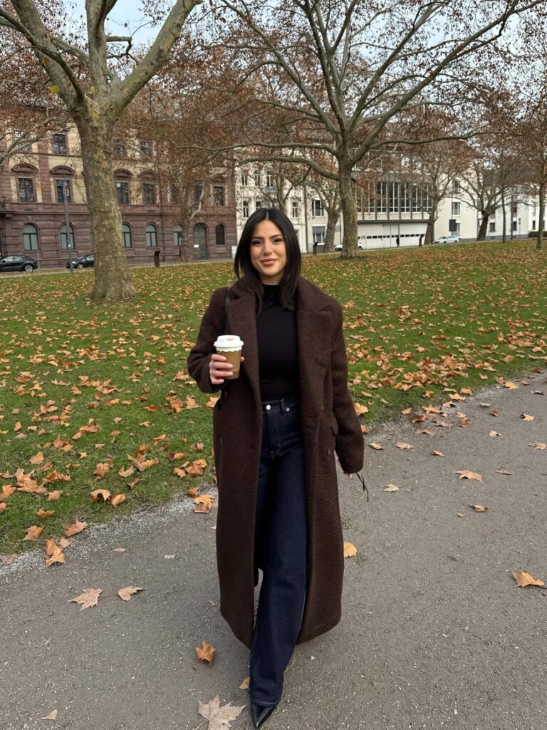 Woman in a brown teddy-textured maxi coat over a black roll-neck with dark wide-leg jeans and black heels in an autumn park